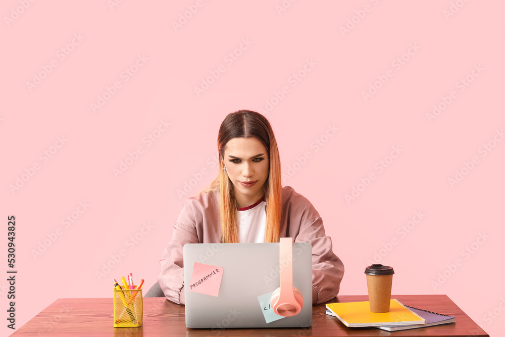 Transgender programmer with laptop at table on pink background Transgender programmer with laptop at table on pink background