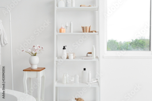 Shelf unit with different bath accessories and vase with flowers on table near white wall © Pixel-Shot