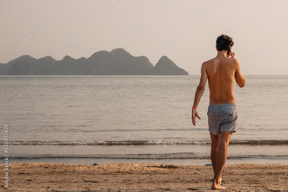 A man in swim shorts talking on the phone on a beach with the ocean and mountains in the background