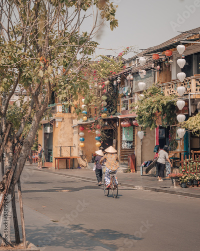 Women wearing Vietnamese conical hats riding a bicycle on a street in Hoi An, colorful houses and lanterns.