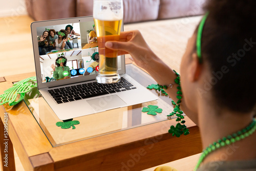 Mixed race woman raising beer glass making st patrick's day video call to friends on laptop at home