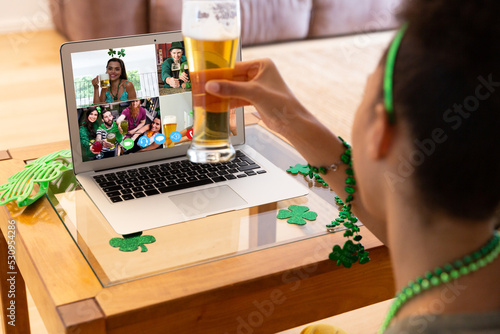 Mixed race woman raising beer glass making st patrick's day video call to friends on laptop at home