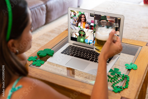 Caucasian woman raising beer glass making st patrick's day video call to friends on laptop at home