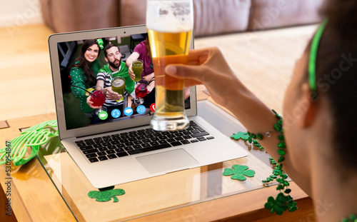 Mixed race woman holding beer having st patrick's day video call with friends on laptop at home