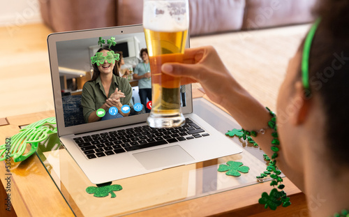Mixed race woman with beer having st patrick's day video call with female friend on laptop at home