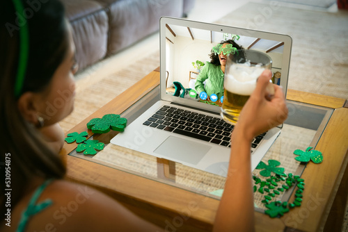 Caucasian woman holding beer having st patrick's day video call with female friend on laptop at home