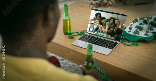 African american man holding beer having st patrick's day video call with friends on laptop at home