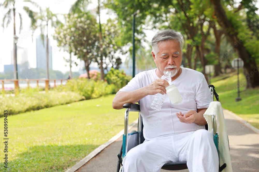 Asian senior man taking medicine with wheelchair at park outdoor. Elderly male drinking water during sunny day Asian senior man taking medicine with wheelchair at park outdoor. Elderly male drinking water during sunny day