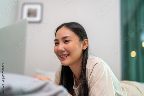 Young Asian woman using laptop computer working freelance work on the bed in bedroom with happiness. Attractive girl enjoy indoors lifestyle with technology online shopping or social media at home.
