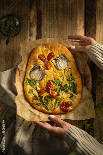 Garden art focaccia bread with vegetables, greens, herbs - beautiful flower composition on homemade freshly baked bread, salty vegan pastry. Wooden table. Copy space. Italian cuisine. Top view