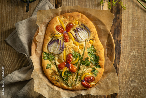 Garden art focaccia bread with vegetables, greens, herbs - beautiful flower composition on homemade freshly baked bread, salty vegan pastry. Wooden table. Copy space. Italian cuisine. Top view photo