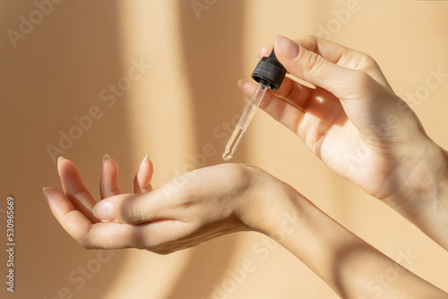 Women's hands hold pipette with cosmetic serum on light brown background, in rays of sunlight. Close-up, copy space. Concept of body care