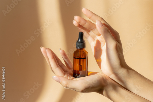 Women's hands hold bottle of amber glass with cosmetic serum on brown background. Mockup of container with dropper lid with collagen in rays of sunlight. Concept of skin care photo