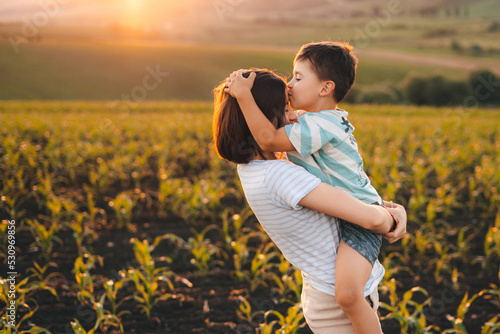 Happy boy kissing his mother on the forehead standing in their cornfield at sunset. They are watching their crops. Happy childhood. Mother's love. Love between