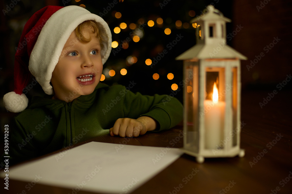 Boy in a Christmas hat writes a letter to Santa Claus by the light of a lantern. Child thinks what to write on the wish list against the background of a Christmas tree decorated with a garland Boy in a Christmas hat writes a letter to Santa Claus by the light of a lantern. Child thinks what to write on the wish list against the background of a Christmas tree decorated with a garland