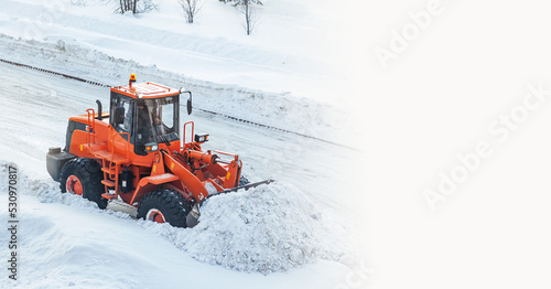 A large orange tractor removes snow from the road and clears the sidewalk. Cleaning and clearing roads in the city from snow in winter. Snow removal after snowfalls and blizzards. banner