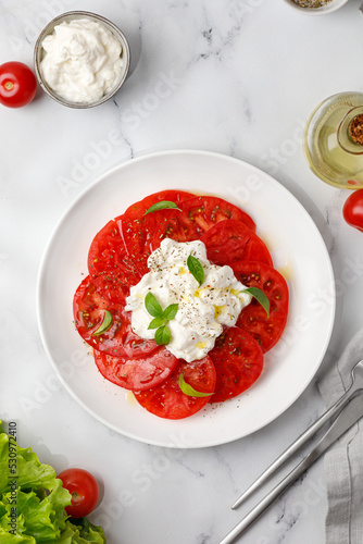 Stracciatella cheese salad on marble background with fork, oil, basil and tomatoes. Top view