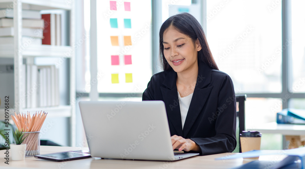 Millennial Asian cheerful successful professional female businesswoman sitting smiling using typing laptop notebook computer at working desk with document folders in company office workstation room Millennial Asian cheerful successful professional female businesswoman sitting smiling using typing laptop notebook computer at working desk with document folders in company office workstation room