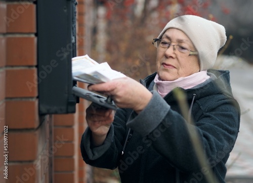 Senor woman opens a mailbox on the street with a key and takes out receipts. Website banner.