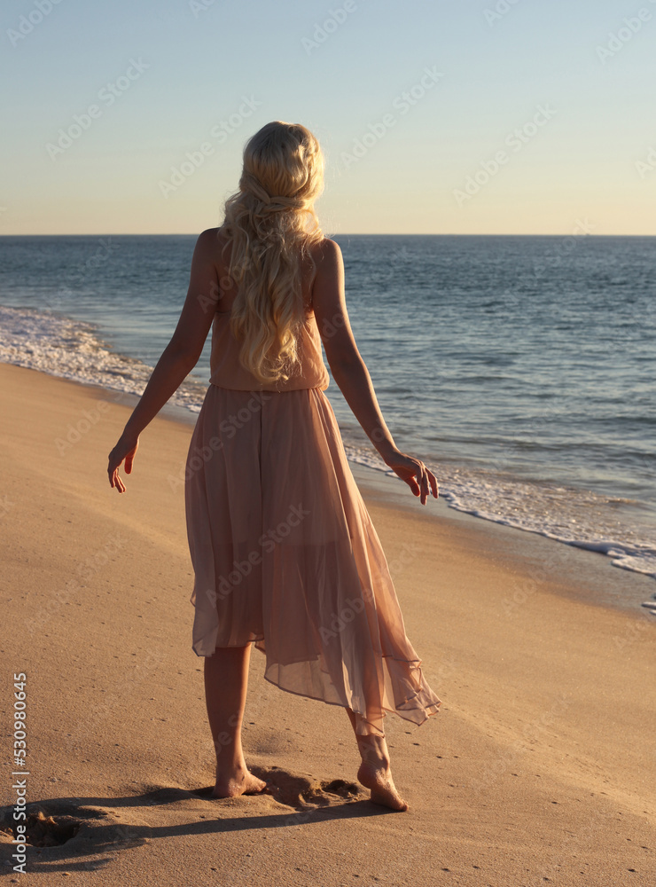 Naklejka premium full length portrait of beautiful young woman with long hair wearing flowing dress, standing pose walking away from the camera. ocean beach background with sunset lighting.