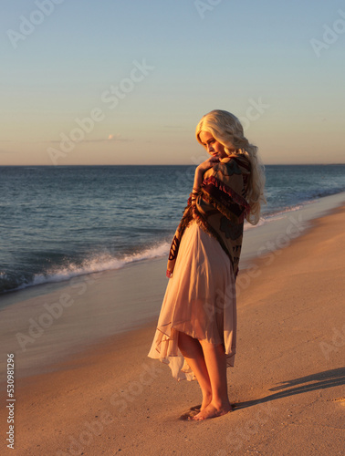 full length portrait of beautiful young woman with long hair wearing flowing dress, standing pose with gestural arms movement . ocean beach background with back lit sunset lighting.