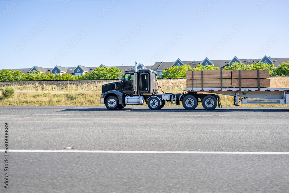 Black big rig semi truck tractor with extended cab transporting lumber cargo on flat bed semi trailer driving on the wide road along the apartments buildings