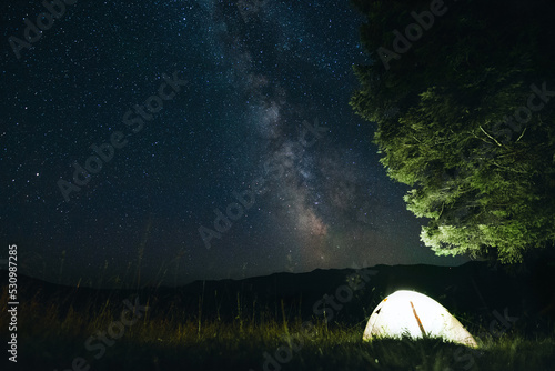 Man tourist near his camp tent at night under a sky full of stars. Orange illuminated tent.