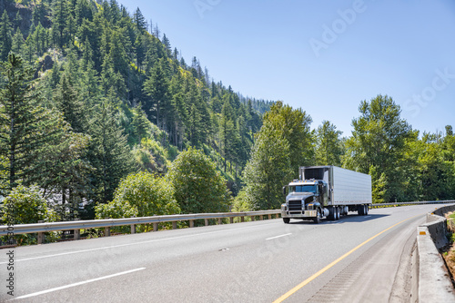 Black low cab profile big rig semi truck transporting cargo in reefer semi trailer moving on the wide one way highway road