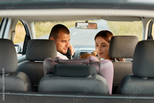 Image of young adult man and woman with little child driving in car, baby daughter sitting in car seat, father and mother looking back at their child.