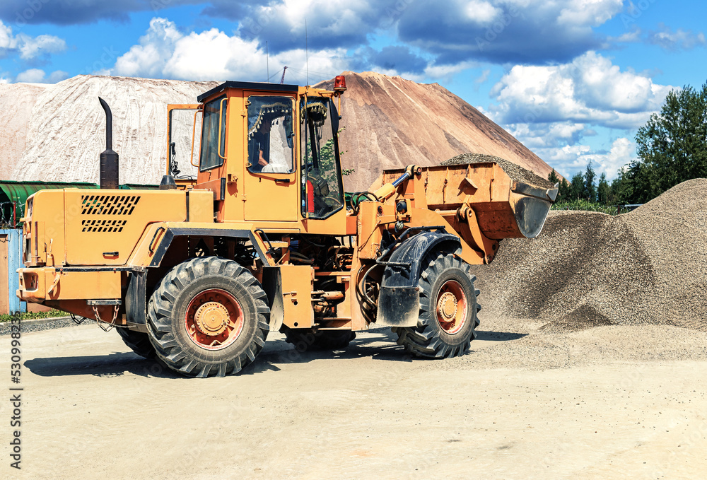 A large front loader transports crushed stone or gravel in a bucket at a construction site or concrete plant. Transportation of bulk materials. Construction equipment. Bulk cargo transportation. A large front loader transports crushed stone or gravel in a bucket at a construction site or concrete plant. Transportation of bulk materials. Construction equipment. Bulk cargo transportation.