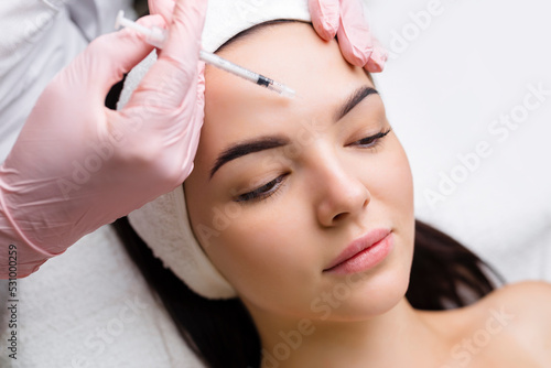 Close up of hands of young cosmetologist injecting botox in female face. She is standing and smiling. The woman is closed her eyes with relaxation photo