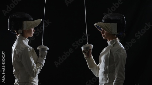 Side view of two young female swordswoman saluting with rapiers. Athletes in white uniforms and protective helmet pose on black studio background. Close up portrait of girls holding epees in hands photo