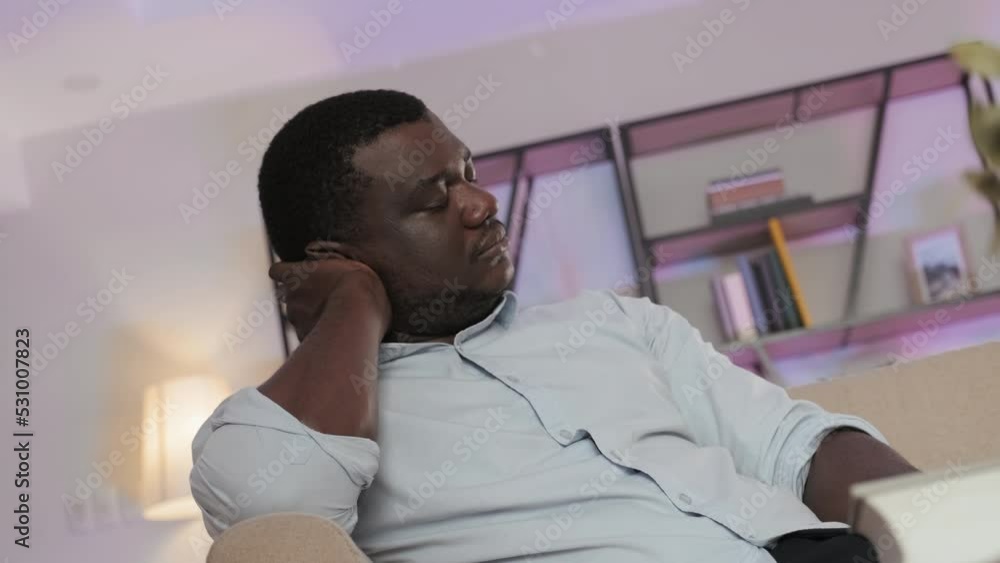 Exhausted Black man in light blue shirt yawning and trying not to sleep after having hard day at work, sitting on sofa in his apartment late at night Exhausted Black man in light blue shirt yawning and trying not to sleep after having hard day at work, sitting on sofa in his apartment late at night