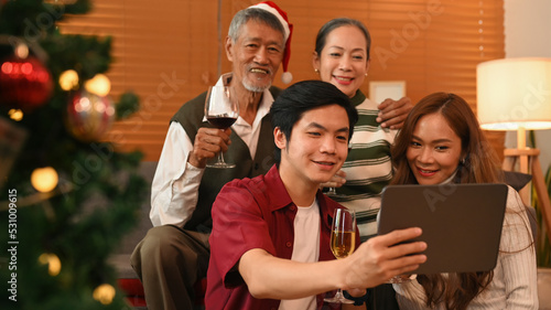 Young couple using digital tablet, taking a selfie with grandparents in living room decorated for celebrating New year and Christmas festive