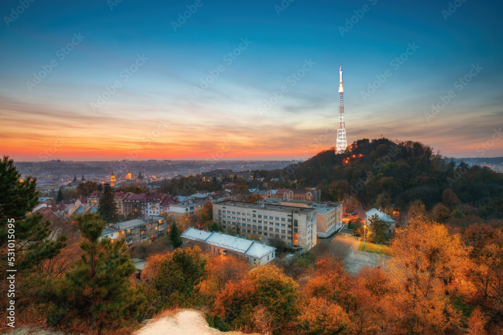 Autumn scenery in Lviv city during sunset, Ukraine. High Castle hill with backlighted TV tower and old quarters in historical city center against colorful sunset sky. Yellowed trees on foreground