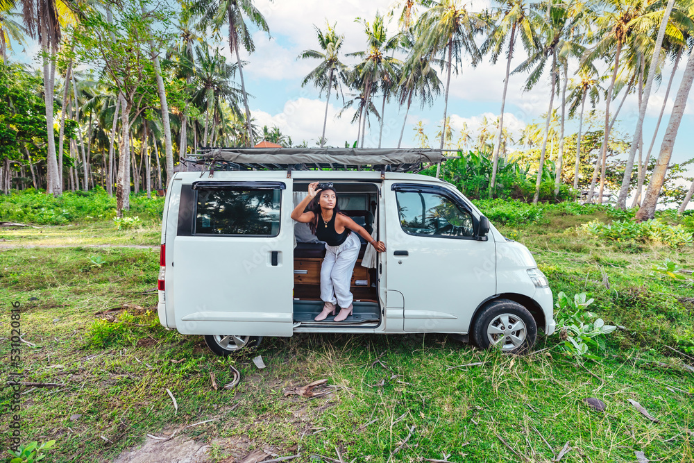 young happy asian girl in a white camper van parked in grass field surrounded by tropical coconut trees in Bali Indonesia on sunny day young happy asian girl in a white camper van parked in grass field surrounded by tropical coconut trees in Bali Indonesia on sunny day