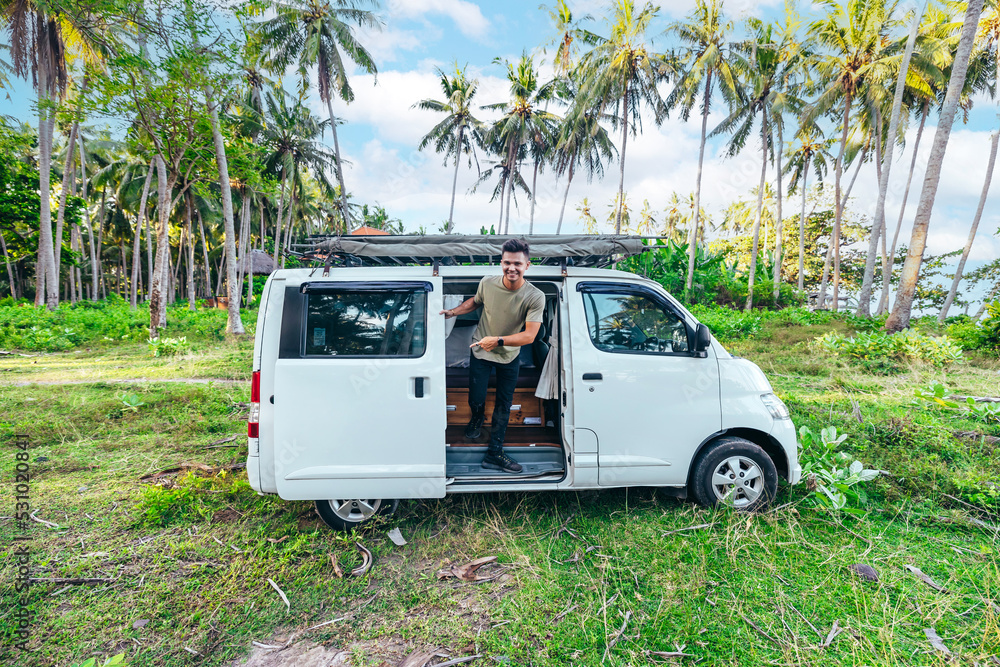 man and white camper van in a grassy tropical field surrounded by coconut trees on sunny day in Bali Indonesia