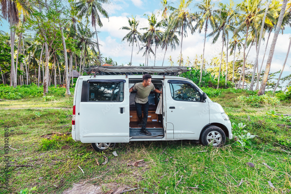 young man looking out of a white camper van parked in a grassy meadow field surrounded by coconut trees in Bali Indonesia on sunny day