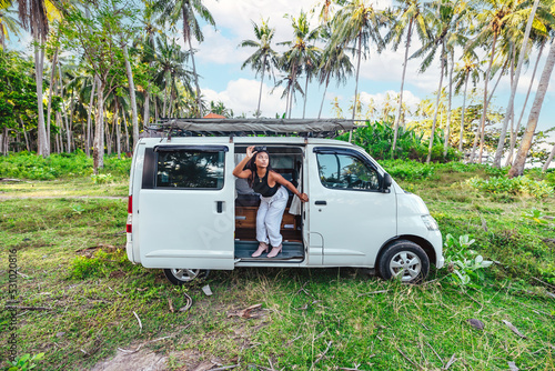 young happy asian girl in a white camper van parked in grass field surrounded by tropical coconut trees in Bali Indonesia on sunny day