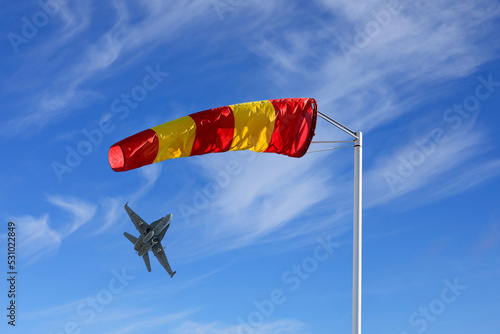 Yellow and red striped windsock or wind cone against clear blue sky, with a combat jet taking off on the background.  photo