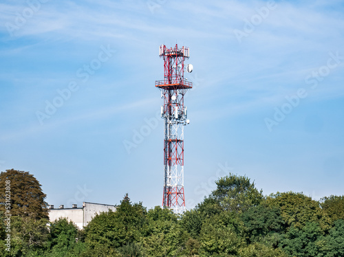 Samotnie wystająca wieża GSM sponad zielonych drzew na tle błękitnego nieba i prawie bezchmurnej pogody