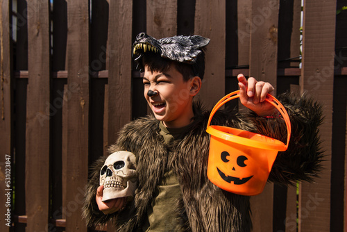 excited asian boy in werewolf costume holding halloween bucket and spooky skull while looking away