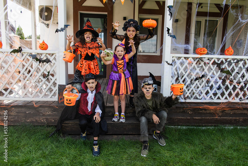 multiethnic kids in halloween costumes grimacing and showing scary gestures near decorated cottage