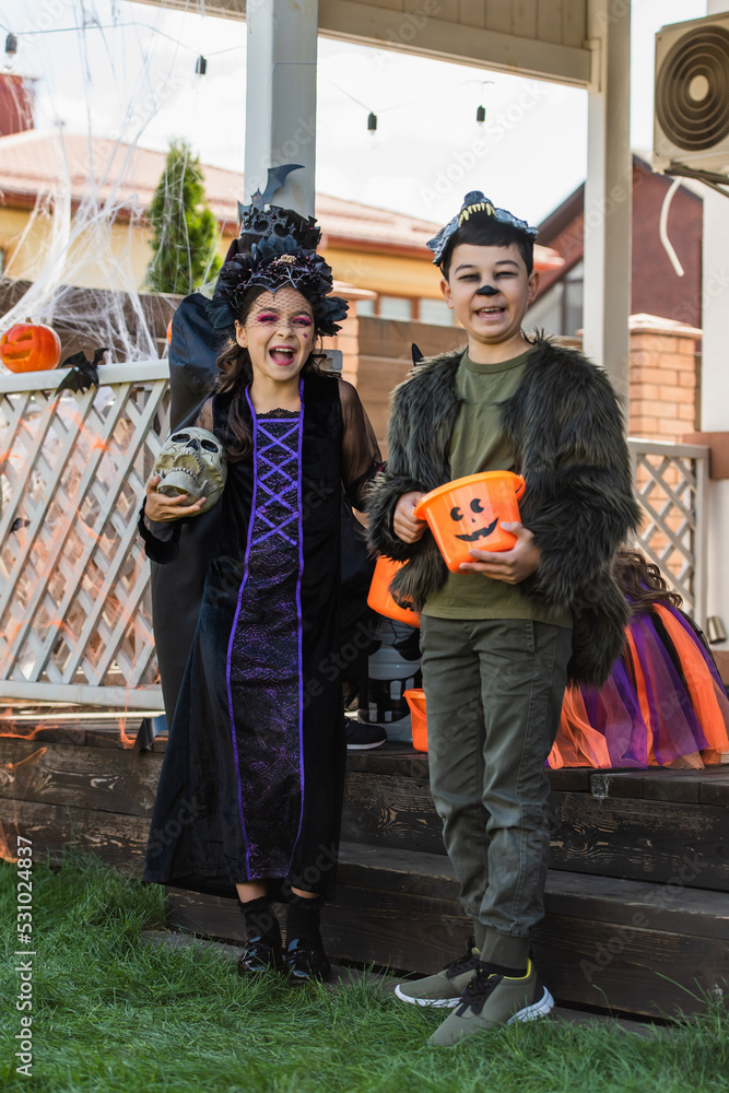 Positive interracial kids in halloween costumes holding skull and bucket on grass in backyard