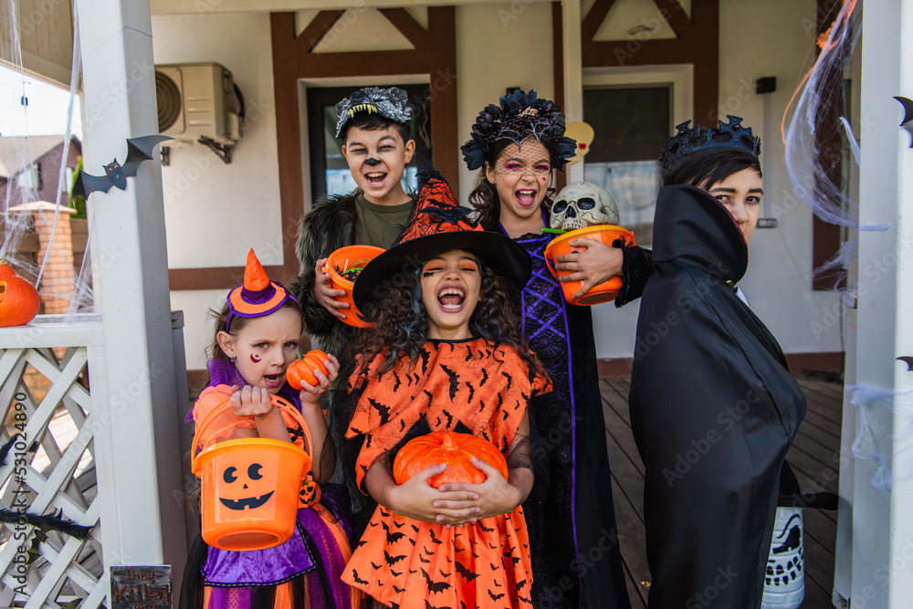 Excited multiethnic children holding buckets with candies near decor during halloween celebration in backyard