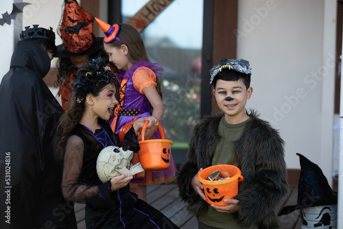 Smiling asian boy in halloween costume holding bucket with candies near excited friend with skull outdoors