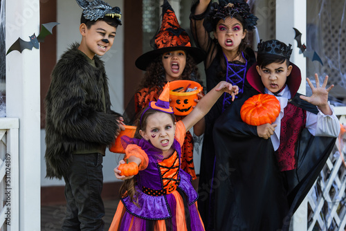 Multiethnic kids in halloween costumes grimacing at camera near house outdoors