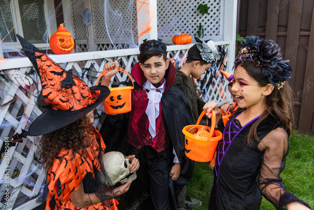 Asian boy in costume looking at camera near cheerful friends with bucket talking during halloween celebration outdoors