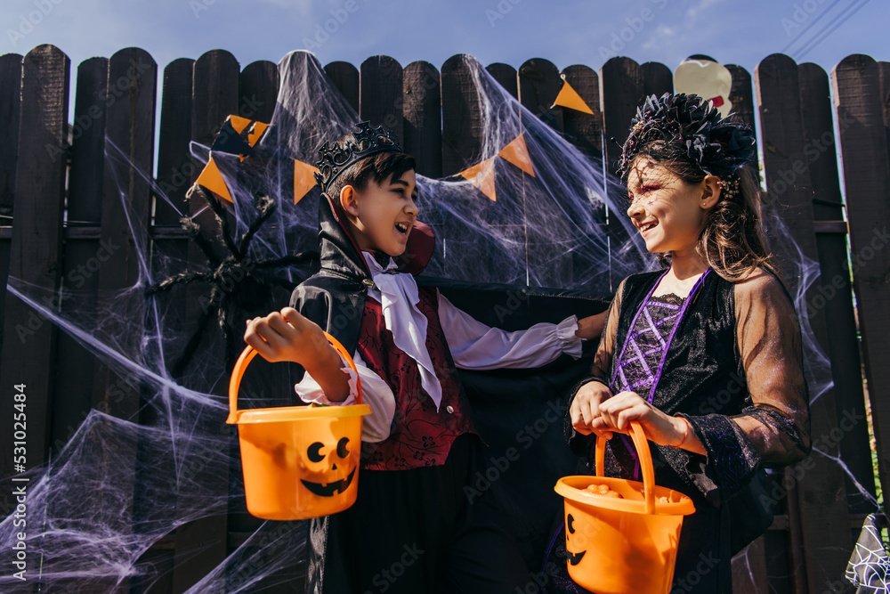 Asian boy in halloween costume holding bucket near smiling friend in backyard