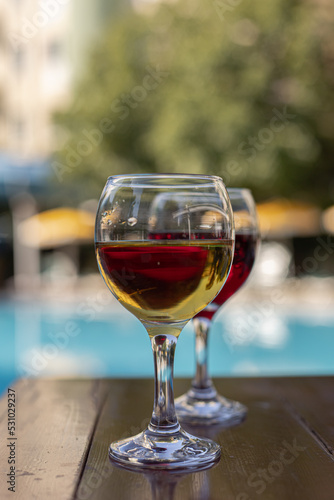 Two glasses of wine on a wooden table next to the swimming pool in summer. Relaxing vacation, holiday and happy weekend concept.